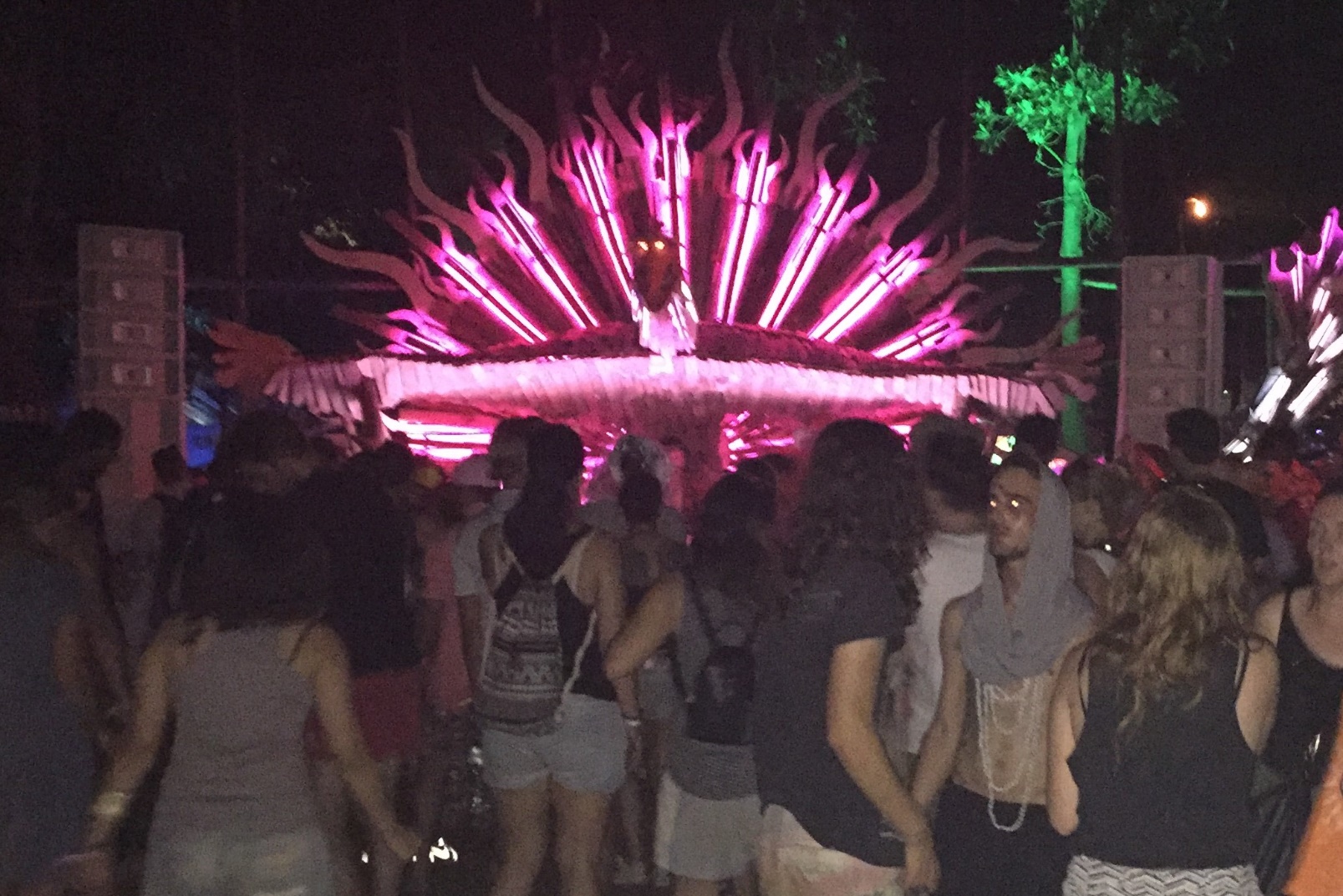 A festival audience faces a wooden stage with tall speaker cabinets on either side, lit at night.