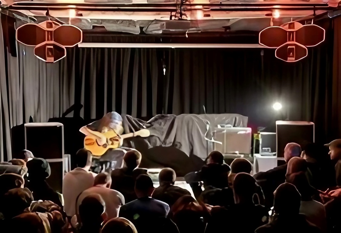 Suspended speaker cabinets above an acoustic guitarist and a seated audience.